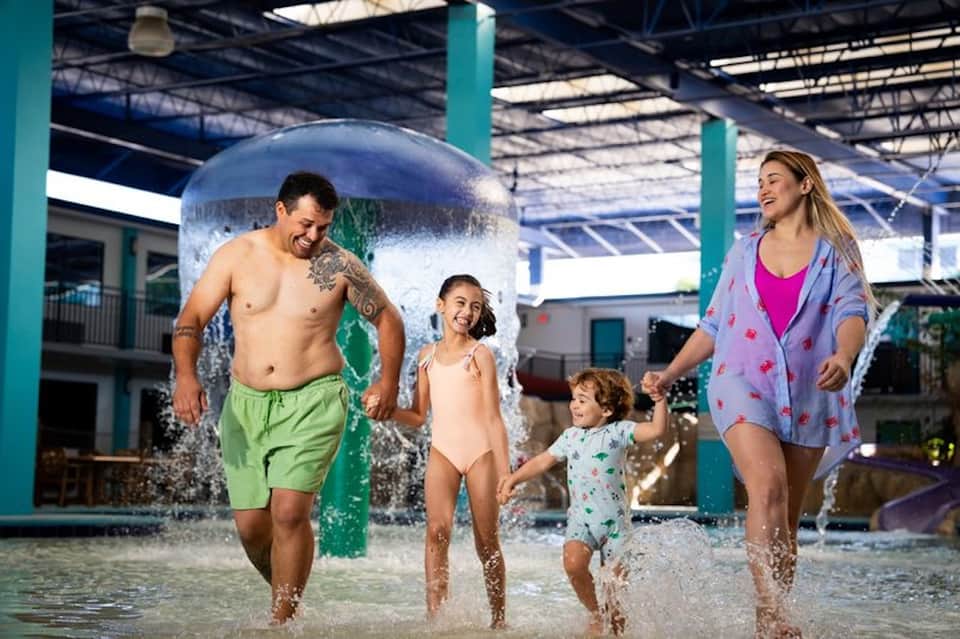 Family enjoying indoor water park with splash pad, turquoise support columns, and tropical setting
