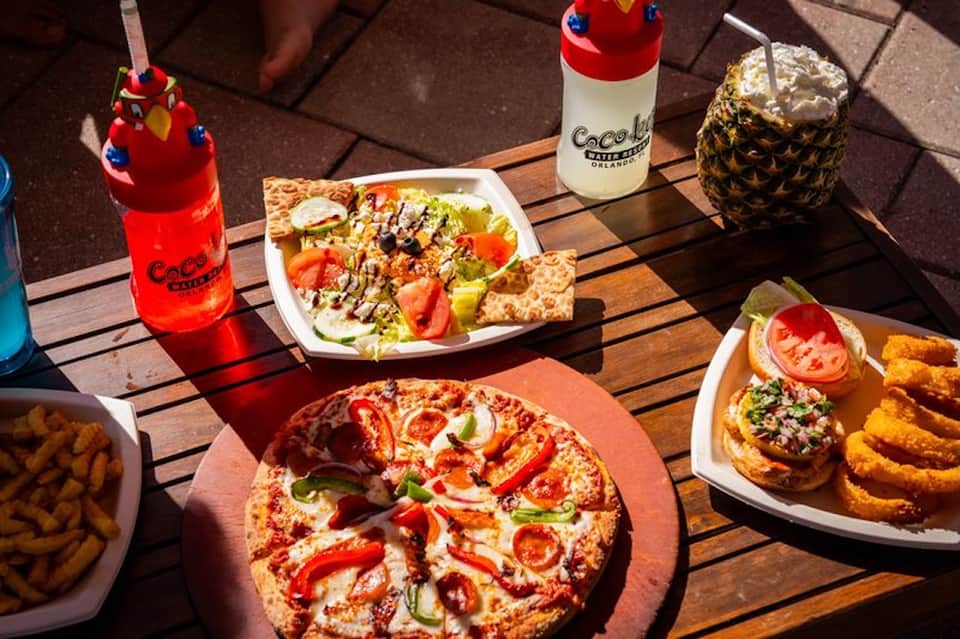 Outdoor patio table with pizza, salad, appetizers, and colorful tropical drinks in sunlight