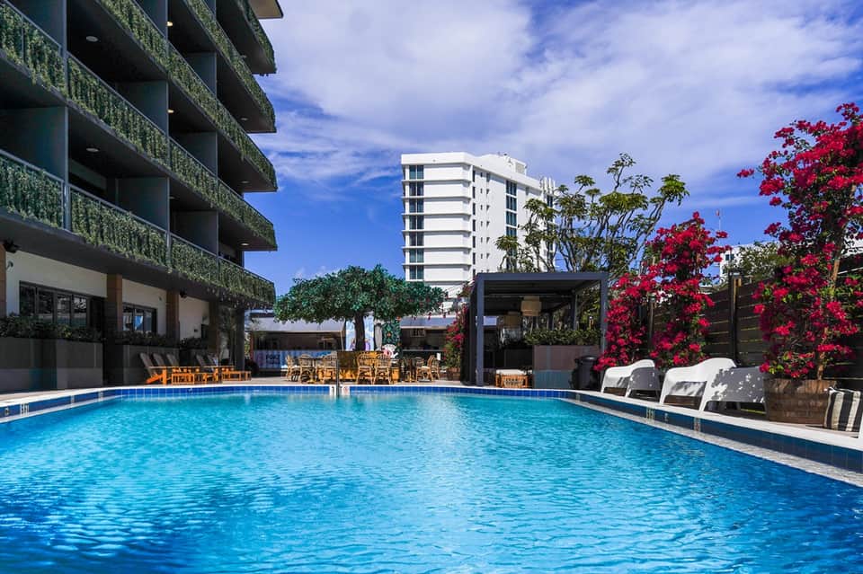 Resort pool area with blue water, lounge chairs, tropical flowers, and multi-story building backdrop