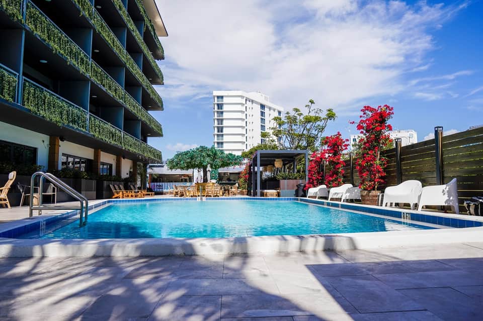 Outdoor swimming pool with white lounge chairs, flowering plants, and modern multi-story building