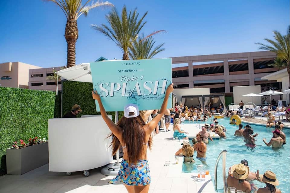 Woman holding turquoise splash sign at busy pool party with crowded swimming pool and palm trees