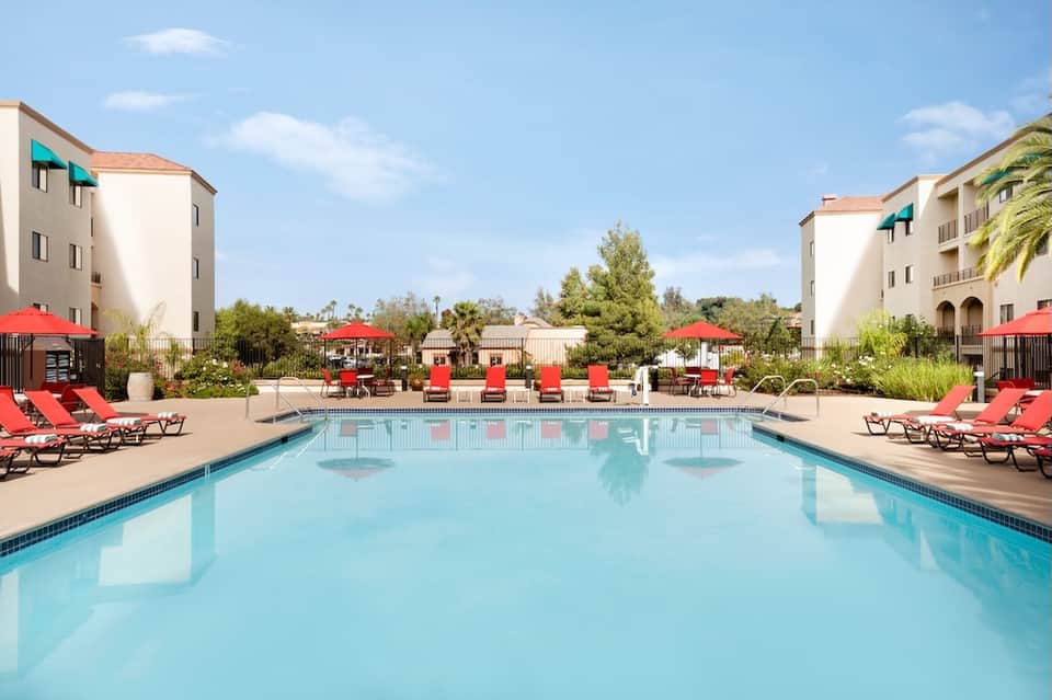 Resort swimming pool with red loungers and umbrellas, surrounded by apartment buildings and landscaping
