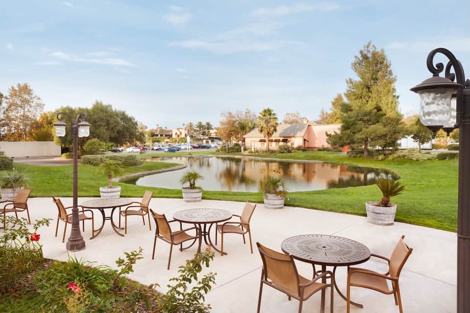Outdoor patio with round tables, chairs, and decorative lamps overlooking pond and residential area