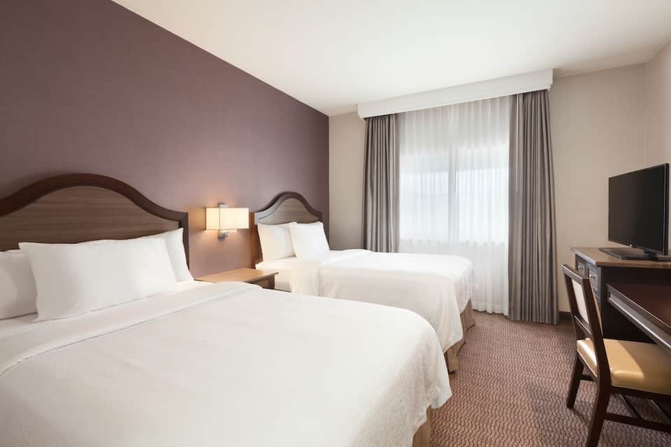 Traditional hotel room with two queen beds, mauve accent wall, and warm wood furnishings