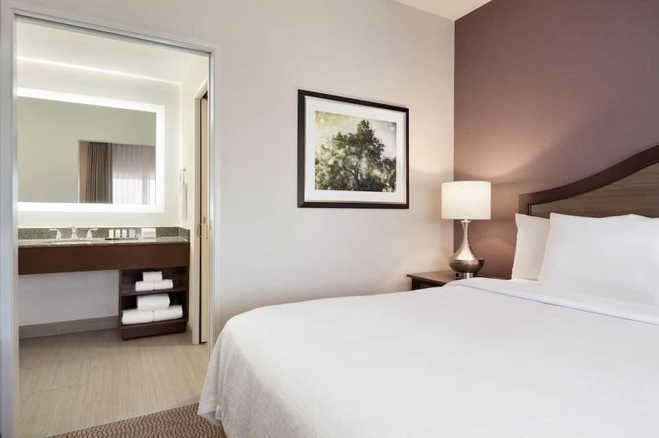 Modern hotel bedroom with king bed, illuminated mirror in ensuite bathroom, warm brown accent wall, natural lighting