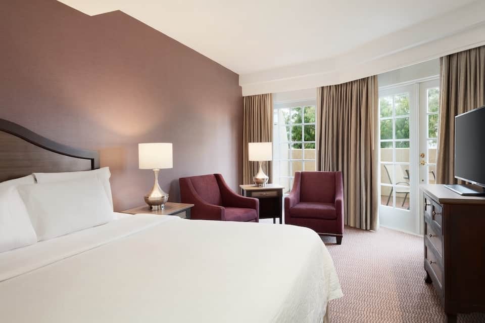 Hotel bedroom with queen bed, burgundy accent chairs, and balcony access