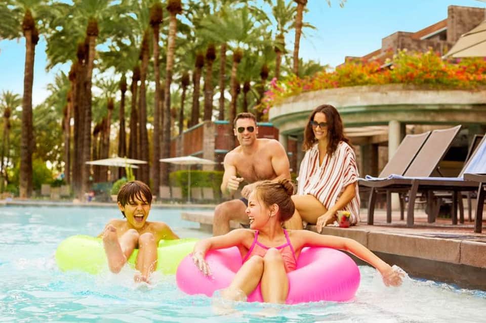 Family playing in resort pool with colorful floats, parents supervising from lounge chairs