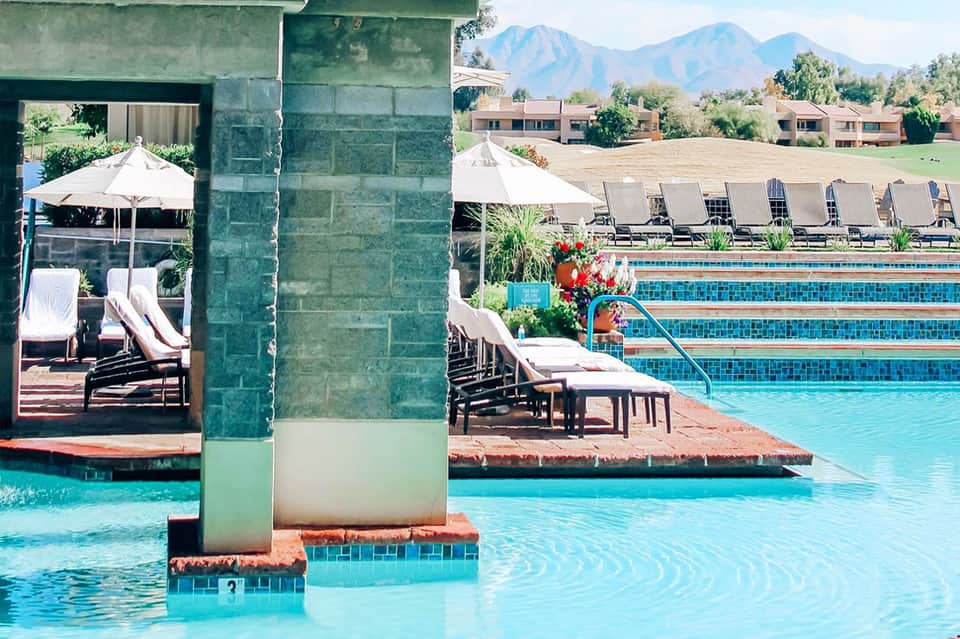 Resort pool with mountain backdrop, loungers, umbrellas, and glass structure entrance