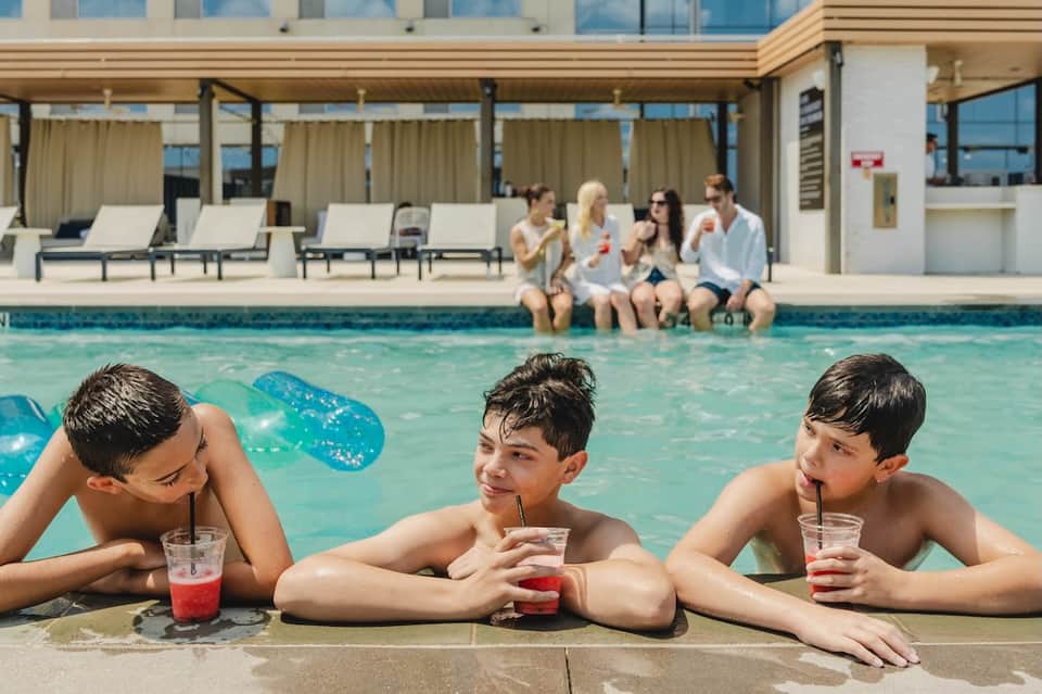 Families enjoying resort pool with children in water, adults sitting poolside, lounge chairs and covered pavilion