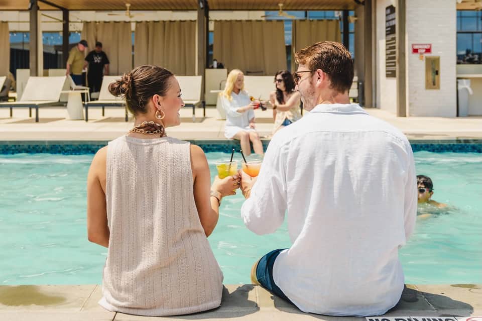 Couple enjoying drinks poolside at resort with other guests visible in the background and covered patio