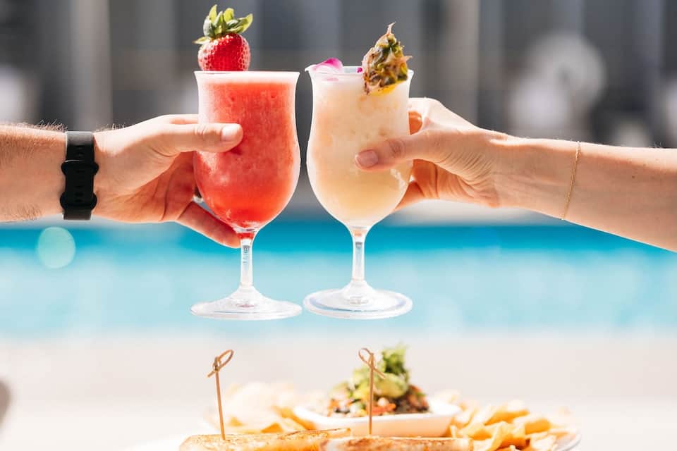 Two hands holding colorful tropical cocktails by pool with appetizer plate below