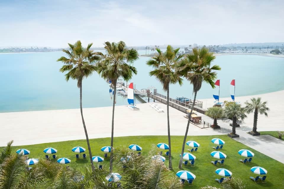 Aerial view of sandy beach with palm trees, wooden pier, blue and white umbrellas, and calm turquoise water