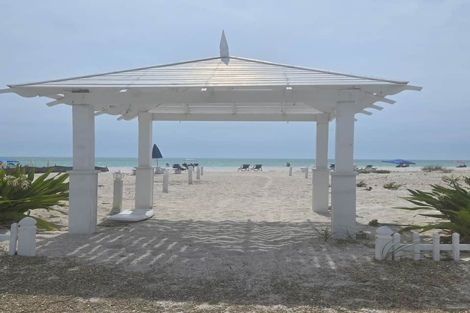 White pergola structure on sandy beach with ocean view and lounge chairs scattered on shore