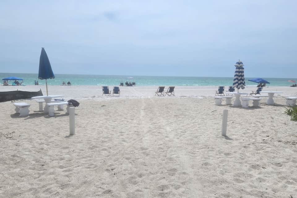 Beach scene with white picnic tables, black umbrellas, lounge chairs, and calm turquoise water