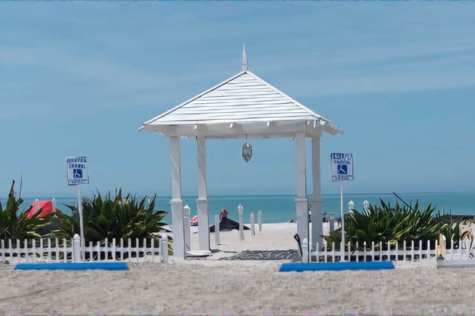 White beachside gazebo on sand with ocean view, white picket fence, and tropical plants