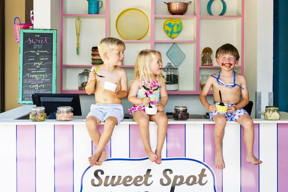 Three young children sitting at ice cream counter with pink shelving and kitchen menu board behind them