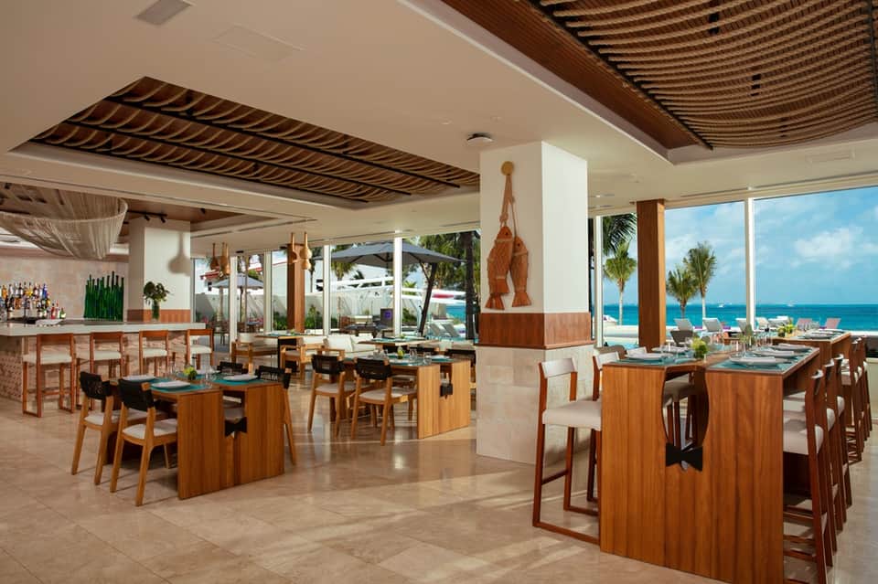 Oceanfront restaurant with wooden tables, bar, ocean views, palm trees, and open-air dining design