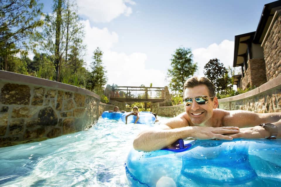 Man relaxing in a narrow resort pool with stone walls, surrounded by green trees and architectural features