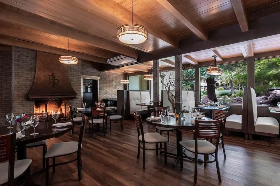 Upscale restaurant interior with exposed brick fireplace, wooden beams, dark furniture, and glass doors