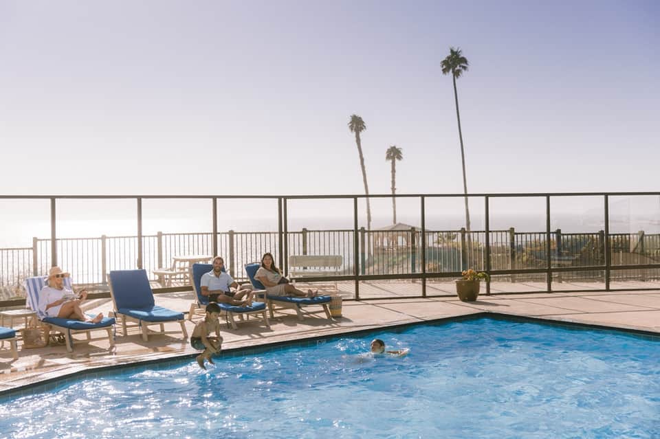 Rooftop pool deck with lounge chairs, palm trees, metal railing, and mountain backdrop at sunrise