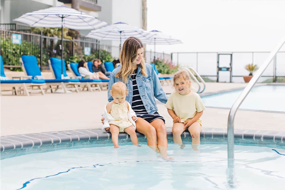 Woman and two toddlers at edge of shallow pool with umbrellas and lounge chairs visible in sunny resort setting