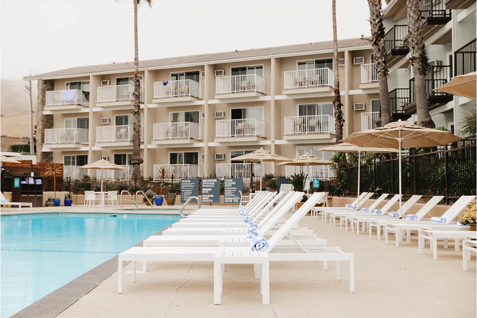Three-story beachfront hotel with cream-colored balconies, palm trees, and white lounge chairs by pool
