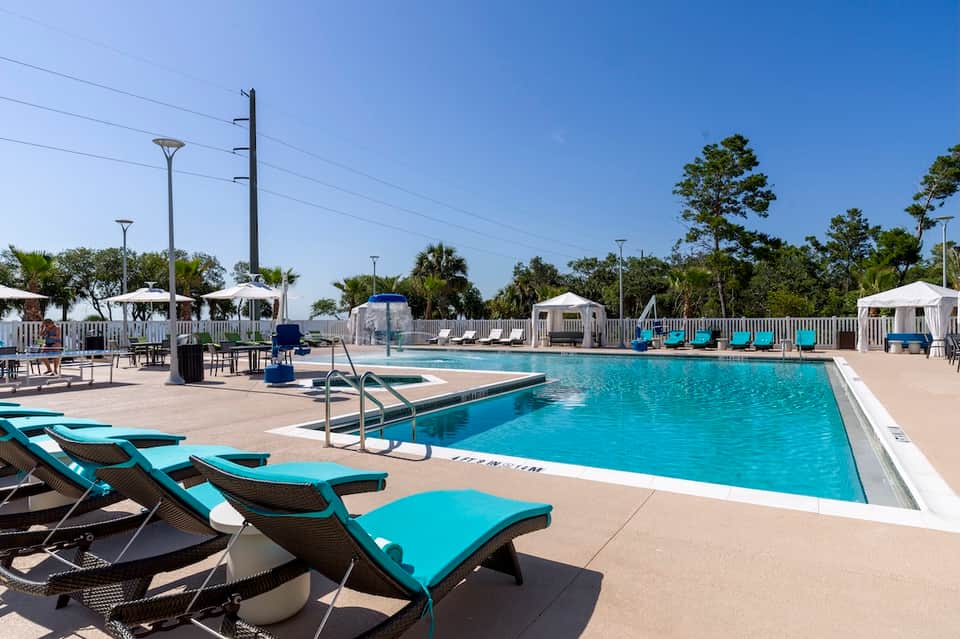 Resort swimming pool with turquoise lounge chairs, white umbrellas, palm trees, and clear blue sky