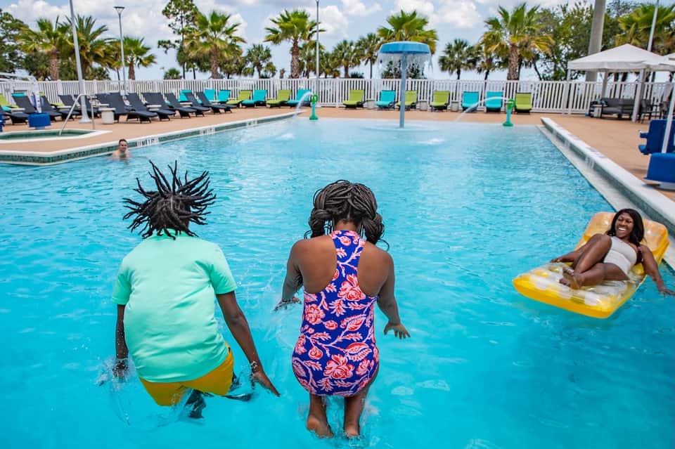 Outdoor pool with swimmers, palm trees, colorful lounge chairs, and white umbrellas