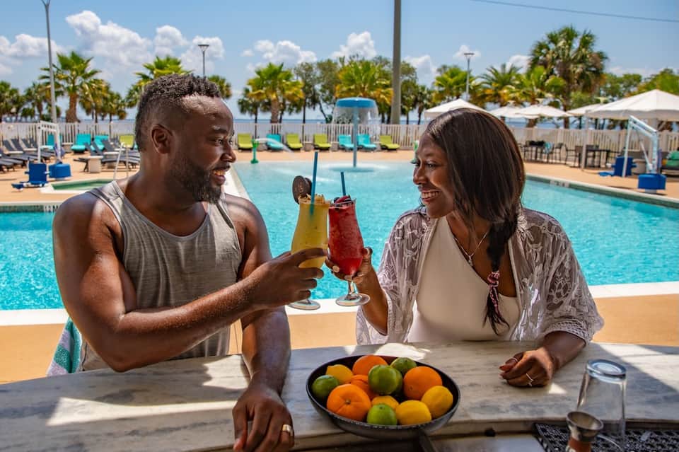 Couple enjoying colorful cocktails poolside with palm trees, beach umbrellas, and lounging areas in background