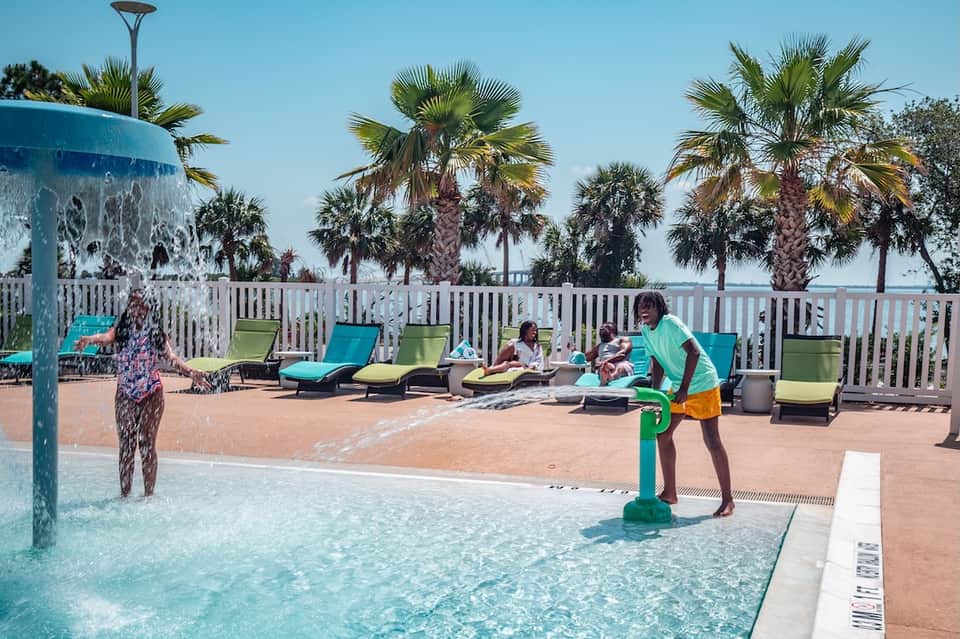 Family enjoying resort pool and splash pad with palm trees and colorful loungers