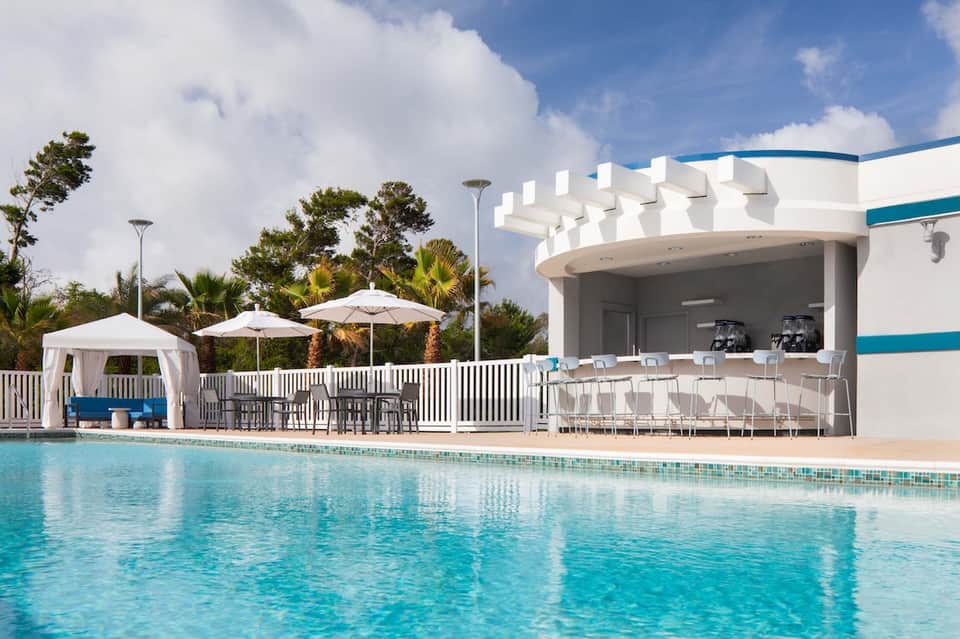 Resort swimming pool with modern white cabana pavilion, lounge seating, and palm trees under blue sky