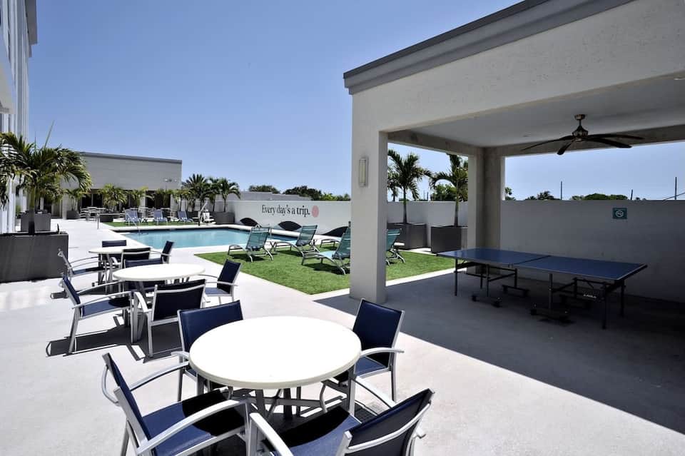 Modern resort pool area with lounge chairs, shaded pavilion, ping-pong table, and palm trees