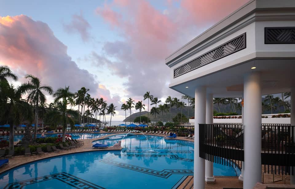Resort pool at dusk with white pavilion, palm trees, lounge chairs, and pink sunset sky