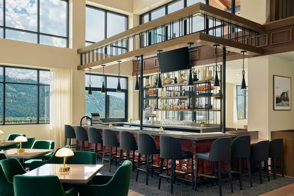 Modern lodge interior with floor-to-ceiling windows, mountain views, green armchairs, and elevated bar