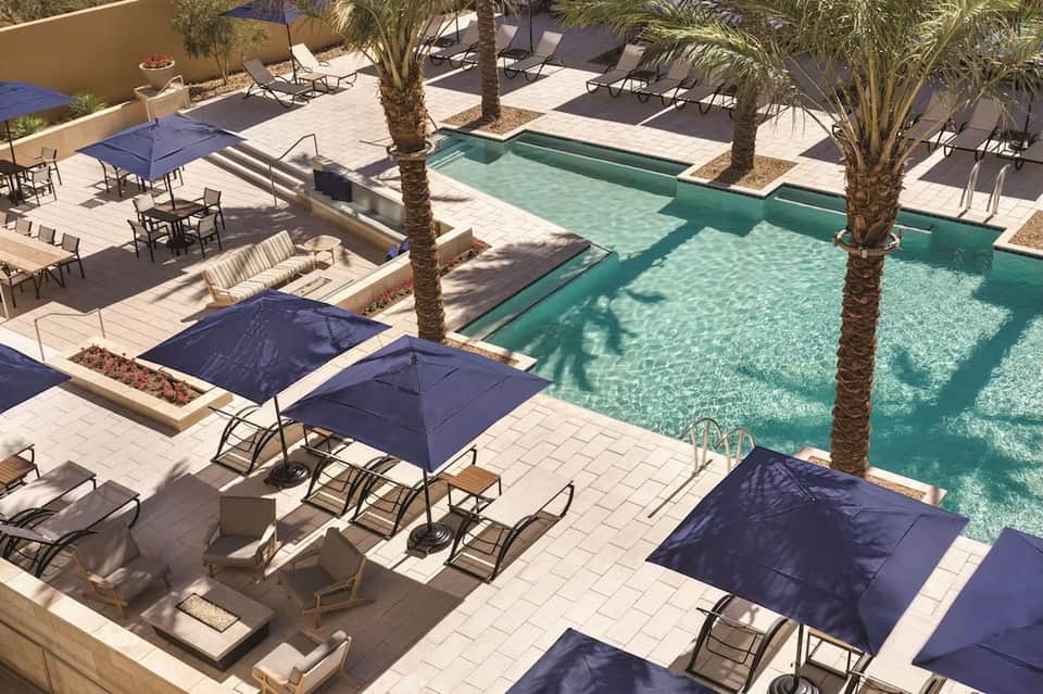 Tropical resort pool surrounded by palm trees, lounge chairs with blue umbrellas on patio deck