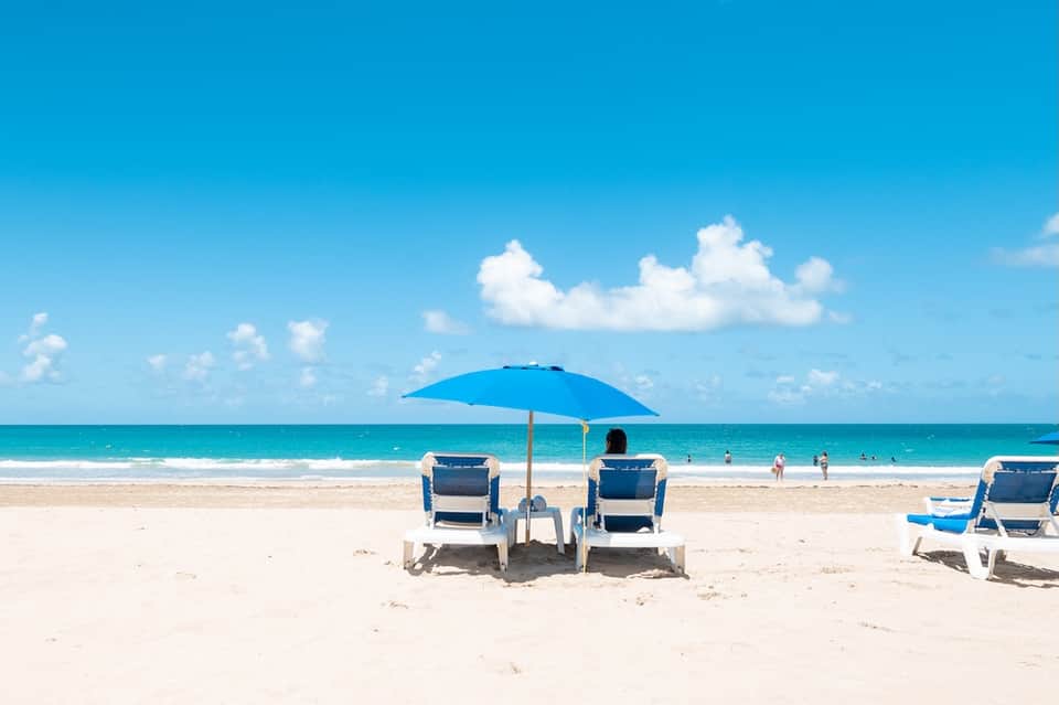 Pristine sandy beach with turquoise ocean, beach chairs under blue umbrella, and swimmers in the water