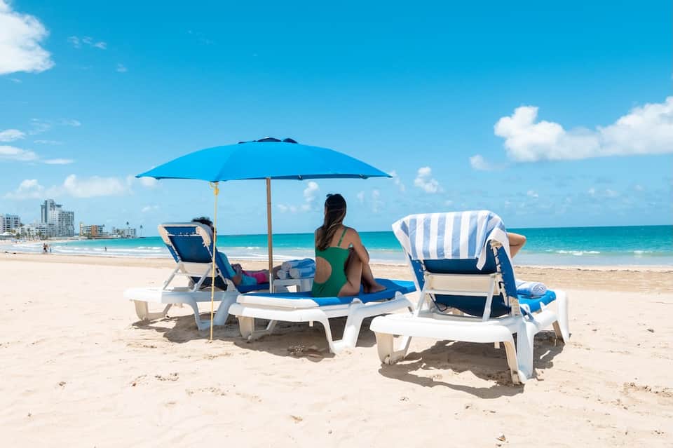 Beachgoers relaxing under blue umbrella with striped lounge chairs on sandy beach with ocean and buildings