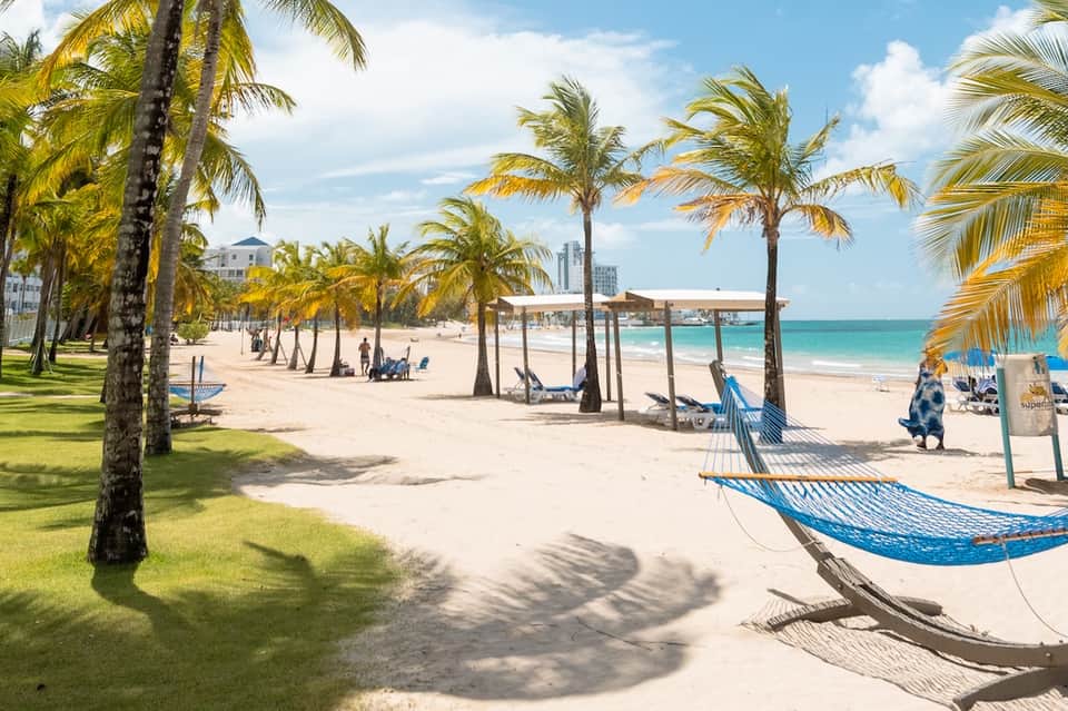 Tropical beach scene with palm trees, hammocks, white sand, turquoise ocean, and buildings along the shoreline