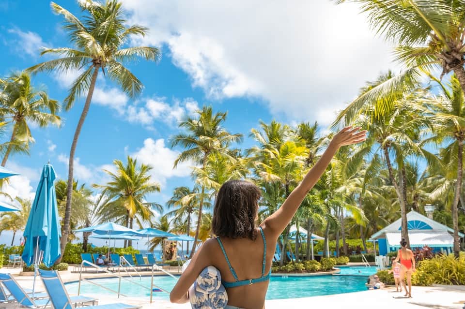 Woman in swimwear at tropical resort pool surrounded by palm trees and blue umbrellas