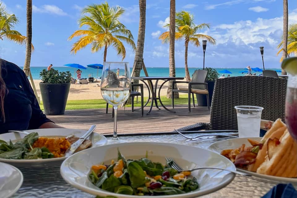Beachfront dining table with salads and drinks overlooking palm trees, ocean, and beach lounge area