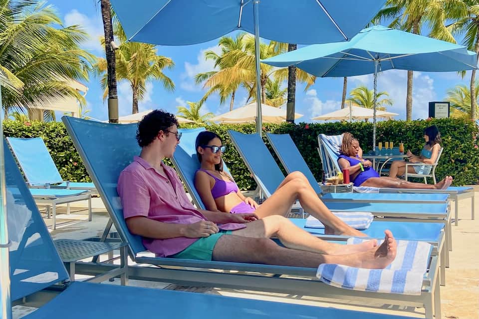 Resort beachfront with guests relaxing in blue lounge chairs under umbrellas with palm trees and blue sky