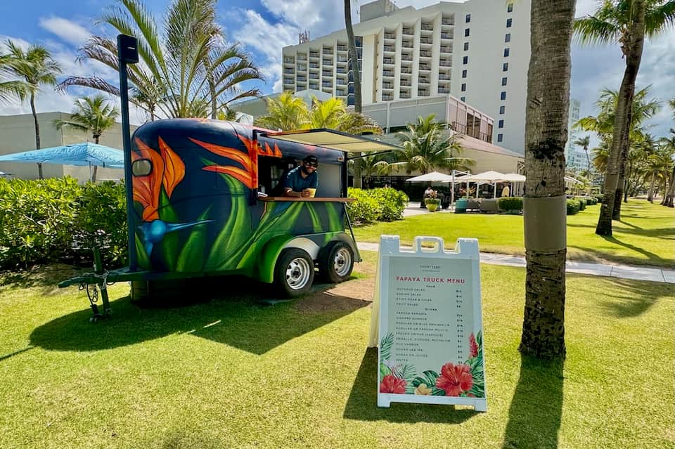 Colorful papaya food truck on resort lawn with palm trees and multi-story hotel building in background