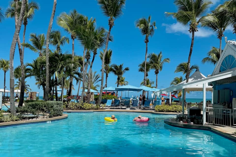 Tropical resort pool with palm trees, cabanas, lounge chairs, and clear turquoise water