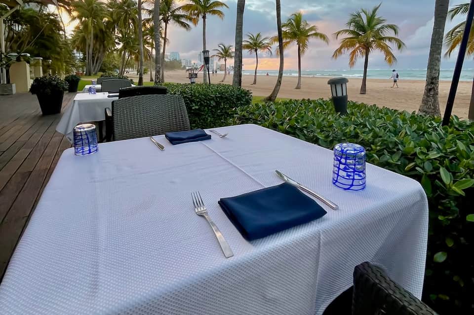 Beachfront dining table set for dinner with ocean, palm trees, and sandy beach visible in background