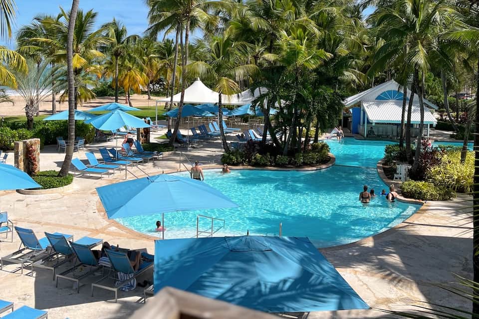 Tropical resort pool with turquoise water, palm trees, blue umbrellas, and sandy beach area