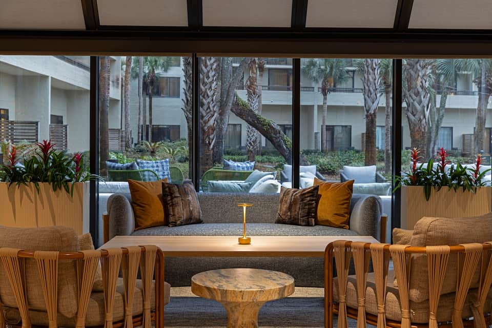 Resort lounge with gray sofa, woven chairs, tropical plants, and large windows overlooking palm-tree courtyard