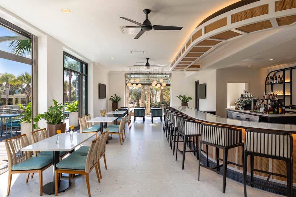 Modern restaurant and bar area with curved wooden ceiling, palm trees visible, dining tables, and outdoor patio