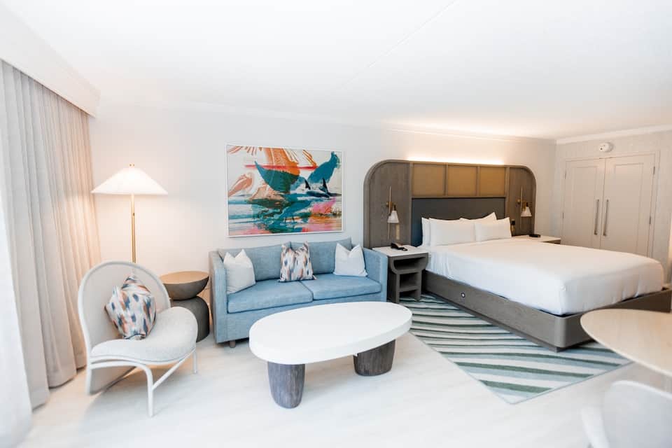 Modern hotel suite with blue sofa, two queen beds, ocean artwork, and balcony access