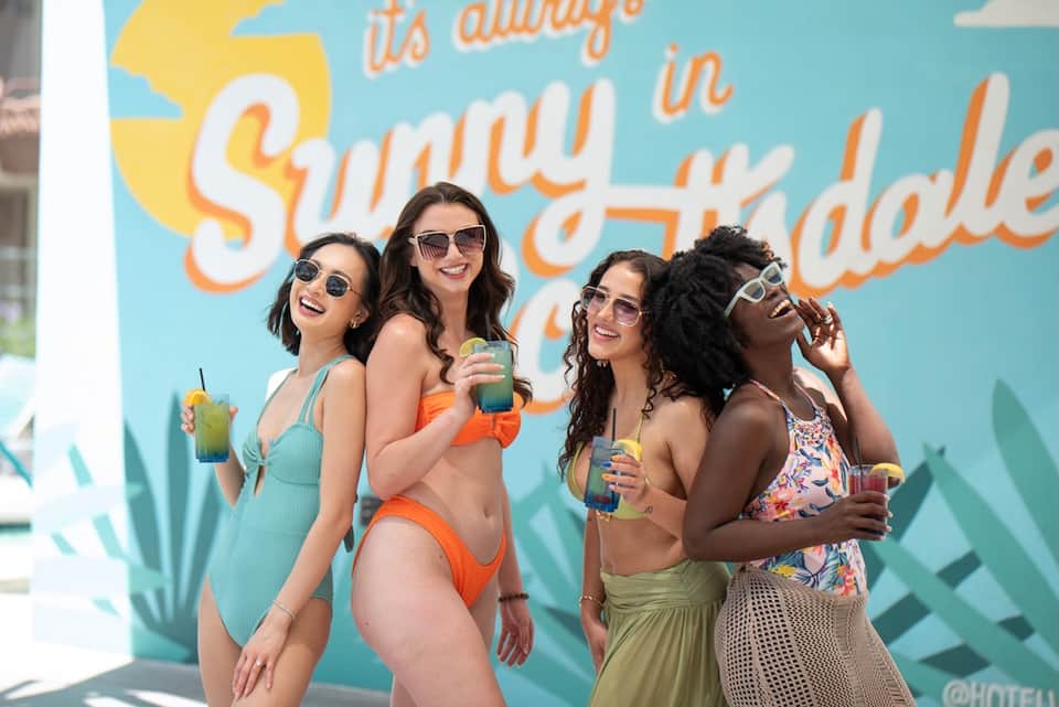 Group of women in swimwear posing by turquoise wall with 'Sunny Hayday' text at pool area