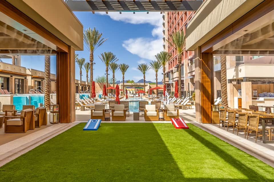 Indoor-outdoor space with green artificial turf, cornhole games, and pool view between buildings